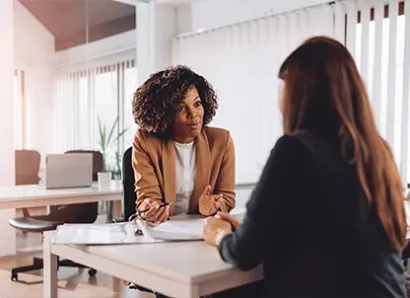 Two women are sitting opposite each other in an office room. One woman shows the other something in a folder.