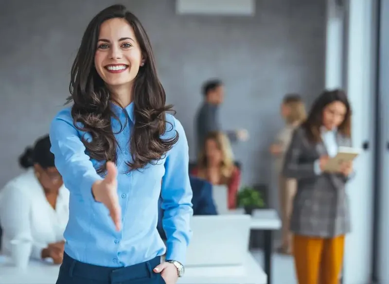 Woman extends her hand in greeting in an office full of employees.
