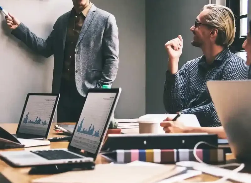 People with open laptops sit together in a meeting room and discuss something on the whiteboard.